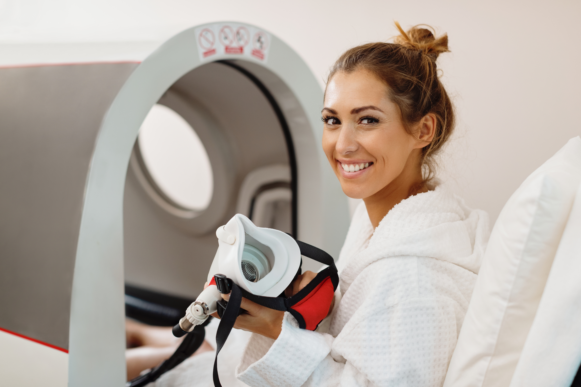 A smiling athlete stands confidently before a hyperbaric oxygen therapy chamber, representing the positive impact of HBOT for athletes on recovery and performance enhancement.